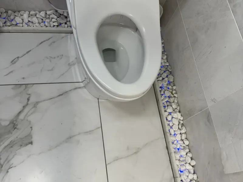 Large-format floor tile cut around a toilet base and closed against a pebble inlay strip